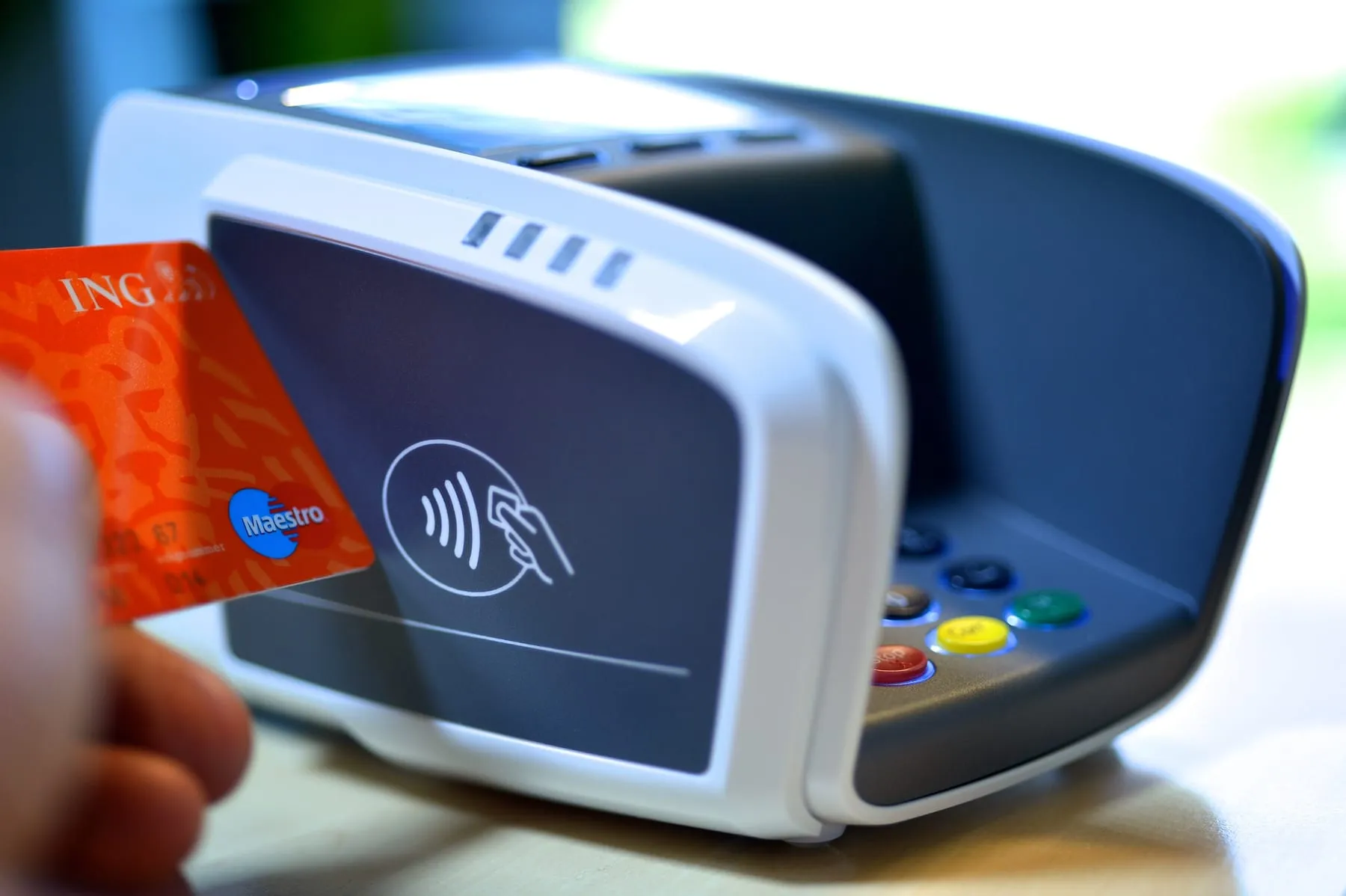 A hand holding an orange contactless bank card up to a white payment terminal, the contactless wave symbol lit on its screen.