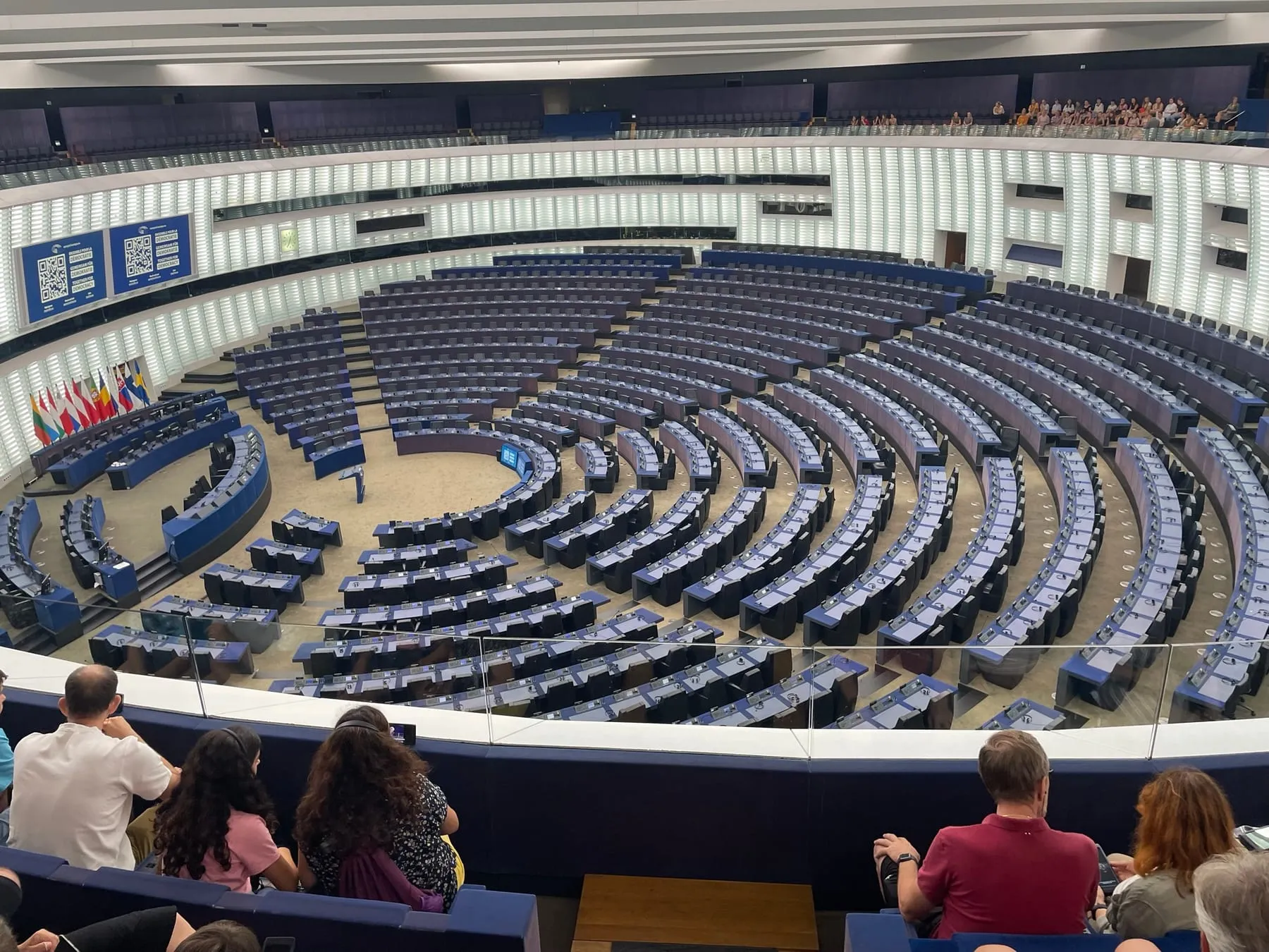 The empty hemicycle of the European Parliament in Strasbourg, a vast circular chamber of pale desks rising in tiers toward a high ceiling.