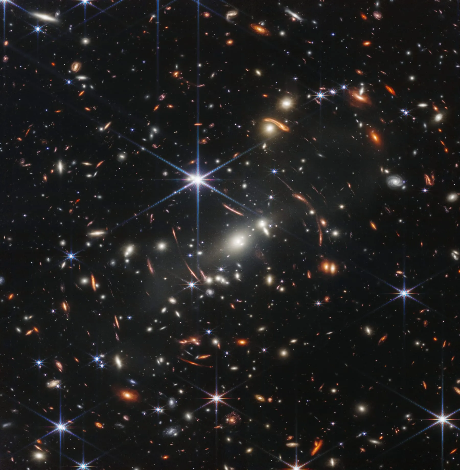 The James Webb Space Telescope's first deep field, thousands of distant galaxies scattered across a black sky, the light of some of them bent by the gravity of a galaxy cluster in front.