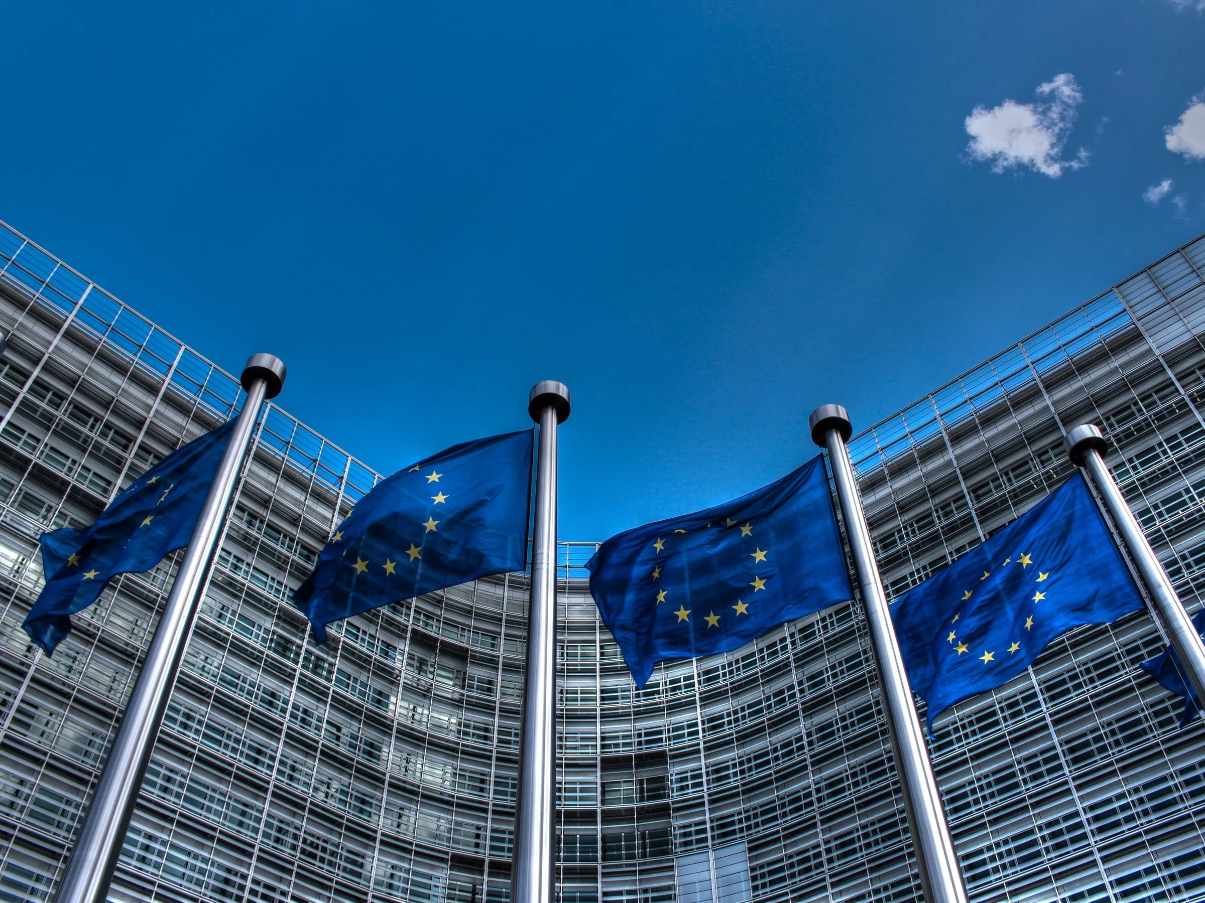 European Union flags flying outside the curved glass facade of the Berlaymont building, the European Commission headquarters in Brussels, against a deep blue sky.