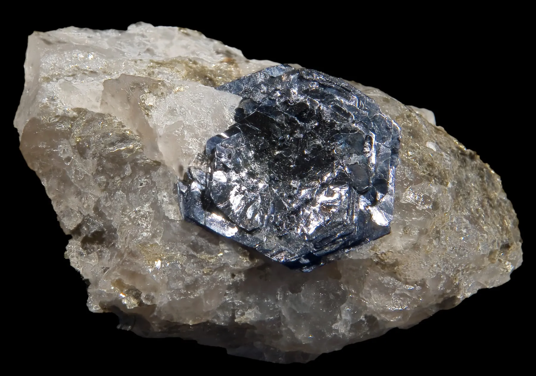 A metallic blue-grey hexagonal crystal of molybdenite, the natural mineral form of molybdenum disulfide, set in white quartz against a black background.